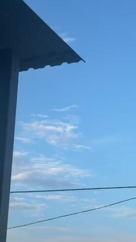 The video shows a view of the sky from under an overhang, likely a roof. The sky is blue with scattered white clouds. In the foreground, the edge of a dark-colored roof is visible at the top of the frame. A vertical white pole is on the left side of the frame. Two dark horizontal lines, possibly wires, stretch across the lower part of the frame. The overall scene suggests an outdoor environment on a clear day.
