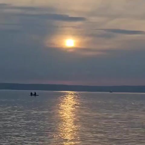 A wide shot captures a tranquil lake scene at sunset. The sun, a bright orb, is partially obscured by clouds, casting a shimmering golden path across the water. In the distance, a sailboat glides across the surface. Closer to the shore, two people are seated on a small boat, likely a canoe or kayak. As the view pans slightly to the right, a sandy beach comes into view. Several people are enjoying the water's edge, including a woman with two children, and another child playing in the shallow water. The overall atmosphere is peaceful and serene, with the warm hues of the setting sun reflecting on the calm water.