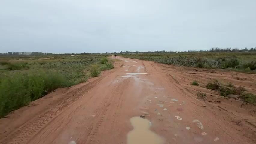 A person walks along a dirt road in a rural landscape under an overcast sky. The road is wet and has puddles, with tire tracks visible. Vegetation lines both sides of the road, consisting of low-lying shrubs and grasses. In the distance, some structures and trees are visible. The camera perspective is from behind the person, following their movement.