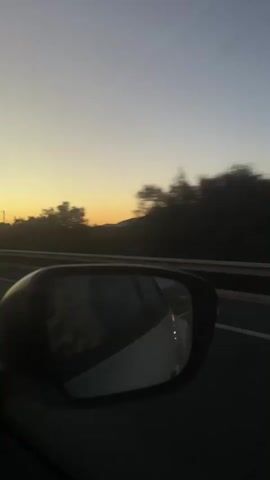 The video shows a view from inside a car, looking out the passenger side window and into the side mirror. The sun is setting, casting a warm orange glow on the horizon. Trees and hills are visible in the distance. The car appears to be moving on a highway. The side mirror reflects the interior of the car and a person sitting in the passenger seat.