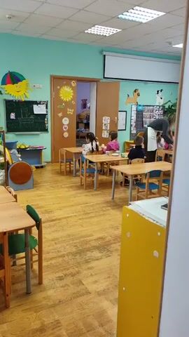 The video depicts a classroom with several children seated at individual wooden tables, each with a small mug and a plate in front of them. A teacher stands nearby, wearing a white apron and tending to the children. The classroom is brightly lit and decorated with various colorful items such as a sun illustration, animal figures, and educational posters. The floor is wooden. An open doorway reveals another room with beds. The children appear to be eating and drinking quietly at their tables.