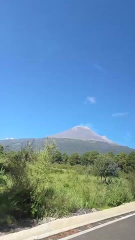 A video shows a view from a moving vehicle. In the foreground, a road is visible with a white curb. Beyond the road, there is lush green grass and various trees. In the background, a large volcano dominates the landscape, with a plume of smoke or ash rising from its peak. The sky is a clear, vibrant blue with a few wispy clouds. The video appears to be filmed during the daytime.