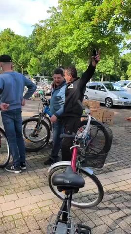The video shows an outdoor market scene. Several people are gathered around bicycles. One man in a black jacket with a white logo on the sleeve is talking and gesturing. Another man in a blue sweater and jeans is standing near a blue bicycle. A third man in a white hoodie and grey shorts is looking at a bicycle. In the background, there are more stalls with items for sale and cars parked. The ground is paved with cobblestones, and there are trees with green foliage in the background. The weather appears to be pleasant, with sunlight filtering through the trees.