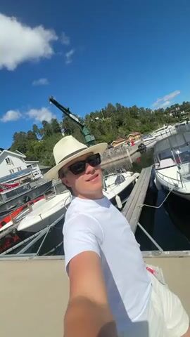 The video shows a young man wearing a white t-shirt, white shorts, a white fedora hat, and sunglasses, taking a selfie at a marina on a sunny day. He is standing on a dock with several boats moored around him. In the background, there are trees, a crane, and more boats. The camera pans to show the marina and the sky. Another shot shows a different person walking away from the camera down the dock.
