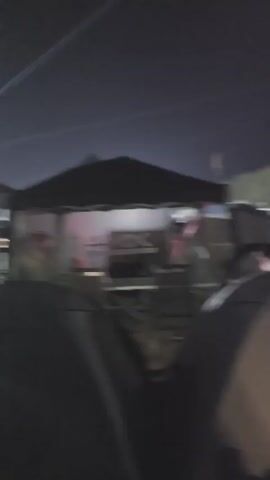 The video shows a blurry night scene, likely an outdoor event or campsite. Streaks of light, possibly from lasers or searchlights, cut across the dark sky. In the foreground, a car's hood is visible, suggesting the footage is from inside a vehicle. The background is filled with indistinct shapes that appear to be tents and possibly other vehicles. There are also lights visible in the distance. The overall impression is of a lively, possibly crowded, nighttime gathering.