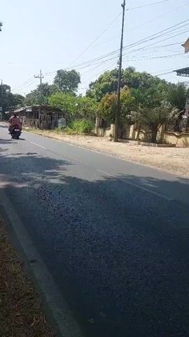 The video shows a road with some vegetation and buildings on the side. There are a few vehicles on the road, including motorcycles and a car. The weather appears to be sunny and warm. In the first few seconds, there are two motorcycles visible, one with two riders, and further down the road, a white car. A second motorcycle with two riders, one in a red shirt, passes by. Another motorcycle with a rider wearing a black hoodie and jeans follows, carrying goods on the back. The rider on this motorcycle appears to be wearing a mask. The motorcycle is also equipped with a pink cargo box and several bags of items on its rear rack. The scene suggests a typical rural or suburban roadside environment on a bright day.
