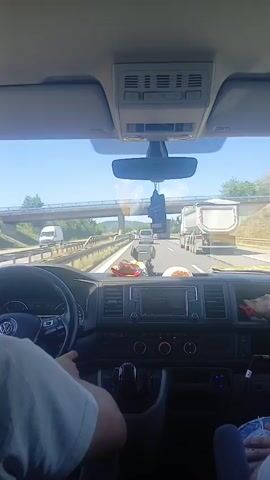 The video shows the interior of a vehicle driving on a highway. A person's hands are visible on the steering wheel and gear shift. The dashboard and windshield are in focus. Outside the windshield, other vehicles, including trucks, are visible on the highway. A bridge and trees line the side of the highway. The sky is blue and appears to be a sunny day. There is an air freshener hanging from the rearview mirror.