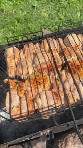 The video begins with a close-up shot of chicken pieces cooking on a grill. The chicken appears to be marinated and is browning nicely. The grill is placed outdoors on a patch of grass, with a blurred background of more grass and greenery. The camera then pans out to reveal a backyard scene. The grill, with the chicken still cooking, is now in the foreground. In the background, there is a well-maintained lawn, a garden area with various plants and flowers, a wooden bench, and a rustic fire pit made of stones and wood. A house with a reddish-brown roof is visible behind the trees. The overall impression is of a peaceful, sunny day in a suburban backyard, perfect for a barbecue.