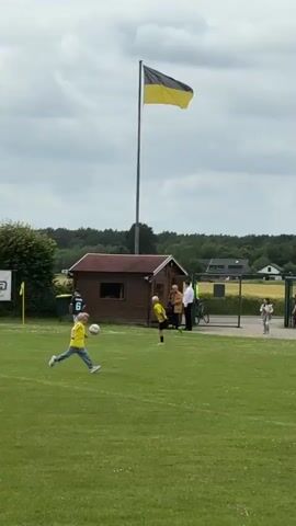 The vertical video captures a soccer field with children playing. In the foreground, a young boy in a yellow shirt and black shorts dribbles a soccer ball. Another boy in a yellow shirt is further away, near a small wooden building. A black and yellow flag waves atop a tall pole behind the building. In the background, people are gathered near a soccer goal, and some individuals walk by, including a woman in a long red dress. The field is green, and the sky is overcast.