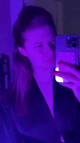 A young woman with light skin and long, light brown hair in a loose ponytail takes a selfie in a room with fluctuating colored lighting. She is wearing a dark, satin-like robe or shirt. Her nails are painted red. The lighting changes from blue to purple to a neutral tone. The woman is holding a smartphone with a glittery case and a pink, textured grip on the back. The phone appears to be an iPhone with multiple camera lenses. The background is slightly blurred, revealing a shelf with various items on it.  There is a white door frame visible behind the woman. She looks directly at the camera with varying expressions, including a slight smile.