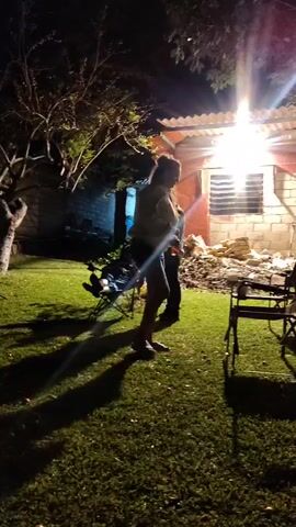 Here's a concise description of the scene:

The scene is a backyard at night, illuminated by a bright overhead light attached to a brick building with a corrugated roof. The yard is covered in grass, casting long shadows. A person is sitting in a folding lawn chair to the left, while another person, wearing shorts and a light-colored shirt, stands near several empty folding chairs. The standing person appears to be talking or looking toward the building. There are also trees and a partial view of another building in the background. The overall atmosphere seems relaxed and casual.