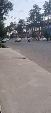 The video captures a street scene in Ismoili Somoni, Tajikistan. A white SUV is the primary subject, driving down a paved road. A person is walking across the street in front of the SUV. The scene is set in a town or city, with buildings and trees lining the street. The time of day appears to be daytime, possibly late afternoon, given the lighting. The weather seems overcast. Visible text includes a license plate on the white SUV, which reads "B 8174 TT". The overall atmosphere is calm and ordinary, depicting a typical urban street scene.