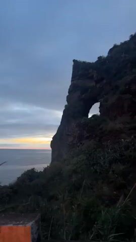 The camera pans from a blurry view of a rocky cliff with a hole in it to a closer view of an old stone bridge with a steep, winding staircase made of stones. The environment is rocky and green, with a cloudy sky above. The bridge appears to be lit from below, highlighting its stone construction. There are no people visible in the video. The focus is on the landscape and the architectural details of the stone bridge and staircase.