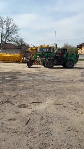 A green heavy-duty vehicle, appearing to be a telehandler or compact wheel loader with a front-mounted bucket, moves across a dirt and gravel lot. A person in a red shirt is visible operating the vehicle from inside the cab. In the background, a larger yellow and green municipal-style truck is parked, featuring equipment often used for road maintenance or snow removal. Partially visible Cyrillic text on the side of this truck reads "КОМПАН." (likely "Компания" meaning "Company") and "СНЕГОРИН" (meaning "snowplow" or "snow removal machine"), confirming its purpose.

To the left of the background truck, several bright yellow metal attachments or containers are stored, consistent with snowplows or salt spreaders. The ground is unpaved and uneven, with several puddles of water reflecting the clear sky. Bare trees stand in the middle-ground, suggesting early spring or late autumn. Houses with roofs are visible in the far background. The scene is brightly lit by the sun under a partly cloudy sky, indicating daytime. The activity takes place in what appears to be an equipment depot or yard.