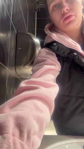 A woman in a black vest over a pink hoodie dries her hands under an electric hand dryer in a public restroom. The bathroom has black tiled walls. The woman activates the dryer and rubs her hands together to dry them. The hand dryer is silver and mounted on the wall. A door is visible behind her.