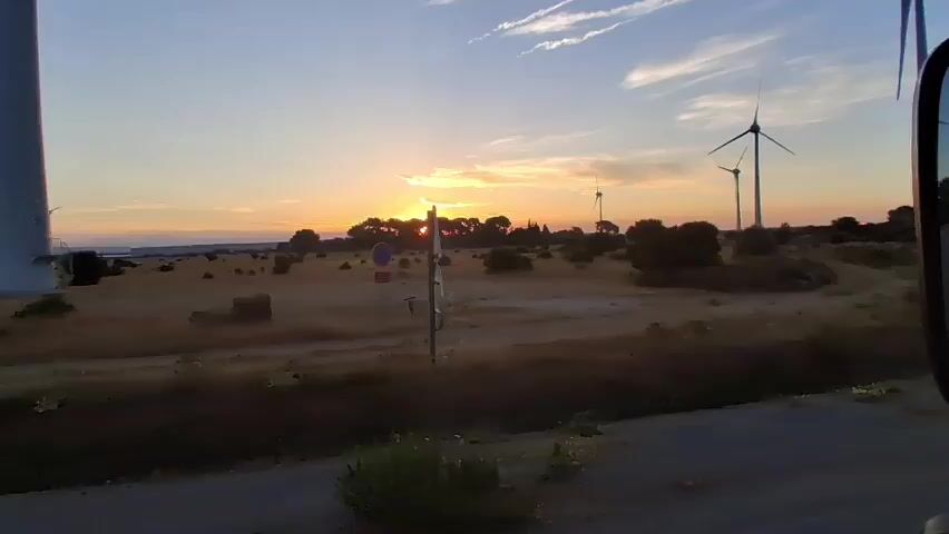 The video shows a landscape with wind turbines at sunset. The sun is low on the horizon, casting an orange glow across the sky. There are several wind turbines visible, with their blades silhouetted against the sunset. The land is covered in dry grass and scattered bushes. A road is visible in the foreground, and a sign with a speed limit of 30 is also present. The video appears to be shot from a moving vehicle, likely a car, as evidenced by the side mirror visible on the right side of the frame. The overall atmosphere is peaceful and serene, with the natural beauty of the sunset complemented by the presence of renewable energy infrastructure.