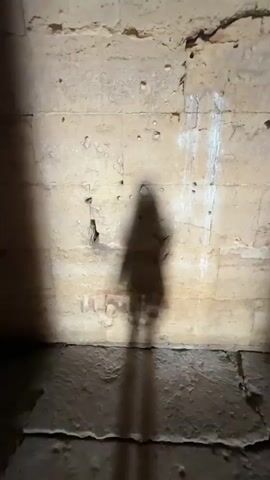 The video shows a dark, stone structure with a rough texture. In the first frame, a shadow of a person is cast against a wall. The camera then pans upwards, revealing concentric circles in the ceiling, suggesting a dome or a circular shaft. A beam of light shines down from above, illuminating the interior. The final frames show a narrow passageway leading towards an opening where sunlight is visible, with the silhouette of a person standing in the distance. The overall impression is that of an ancient or subterranean space.