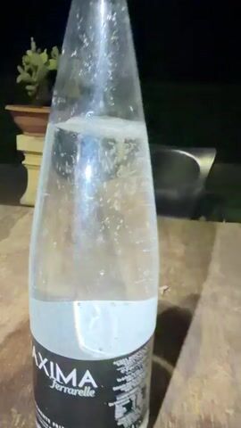 A clear plastic bottle of carbonated water sits on a wooden table outdoors at night. The bottle is labeled "MAXIMA FERRARELLE" and "ACQUA MINERALE FRIZZANTE". A plant in a pot is visible in the background. The lighting suggests artificial illumination.