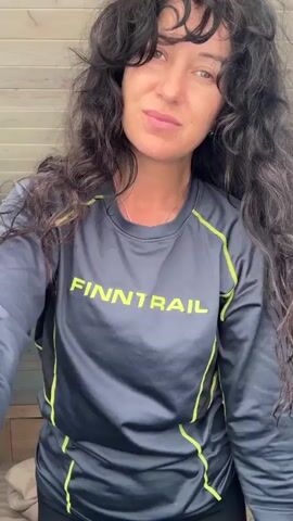 A woman with long, dark, curly hair and bangs is standing in front of a wooden paneled wall. She is wearing a long-sleeved gray shirt with neon green accents and the word "FINNTRAIL" printed in white across the chest. She is also wearing black leggings. In the video, she touches her hair, puts her hands behind her back, and then turns to the side. She smiles and pouts at the camera. The background appears to be a bedroom with a bed and a dresser.