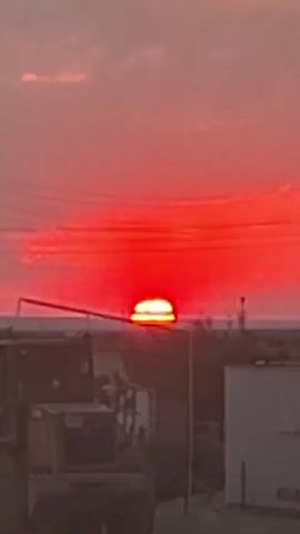 This is a vertical, low-resolution video of a sunset. The sky is a deep red, with some darker clouds. The sun is a bright orange circle low on the horizon, partially obscured by a dark horizontal line. In the foreground, there are some blurry objects that appear to be machinery and buildings. There are also power lines visible in the sky. The video loops.
