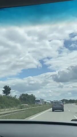 The video shows a view from inside a vehicle looking out at a highway. The sky is filled with white and gray clouds, with patches of blue sky visible. On the highway, there is traffic congestion. Several trucks and cars are visible, some stopped and some moving slowly. The road appears to be a multi-lane highway with barriers separating the lanes. There are trees and vegetation along the side of the road. The overall impression is of a normal day on a highway with traffic delays.