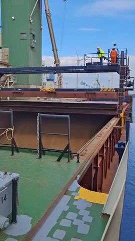 The video shows a scene on the deck of a barge. Two people, one in a yellow safety vest and another in an orange suit, stand on a platform overlooking a large, open cargo hold of the barge. The deck is painted green with some areas marked in yellow and gray. Chains and metal supports are visible along the sides of the hold. In the background, a tall green industrial building stands adjacent to a crane. The sky is blue with light clouds. The barge appears to be docked in an industrial area.