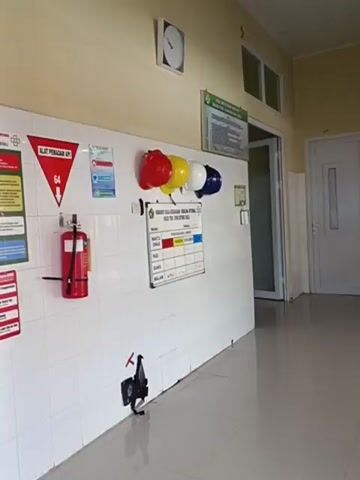 The video shows an interior hallway or room with white tiled walls. On the wall, there are several safety posters and signs, a fire extinguisher, and a whiteboard with colored markers. Several hard hats in red, yellow, and blue are hung on the wall. To the left, there is a door with frosted glass, and to the right, there are two more doors with glass panels. In the reflection of the glass doors, a person can be seen sitting at a desk. There are also several red chairs visible in the foreground and along the wall. The environment appears to be an office or a communal area within a building.