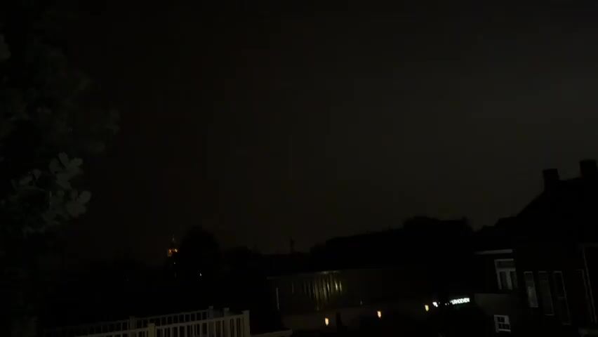 The video is a nighttime shot of a suburban backyard. A large tree is on the left side of the frame, with its leaves silhouetted against a dark sky. In the center of the frame, a faint light is visible in the distance, possibly from a streetlight or a building. The foreground features a white deck railing. The sky is dark and cloudy, suggesting it might be during a thunderstorm or heavy rain. There are no people or active movements visible in the video. The overall impression is one of a quiet, dark evening in a residential area.