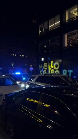 At night, several police cars with flashing lights are parked on a street. The police cars are marked “Boston Police.” The scene is lit by the flashing police lights and the lights from a nearby building, where a sign with the words “HELLO” and another word beginning with the letters “SE” are visible. Several people are walking on the sidewalk.