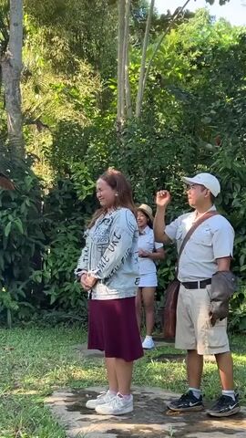 A woman in a denim jacket and maroon skirt stands in a grassy area with trees behind her. An eagle is flying in the air above and to the right of the woman. A man in a khaki uniform with a glove on his left hand stands to the right of the woman, facing her. Another person in a khaki uniform is standing behind the woman. The woman appears to be reacting to the eagle's presence.
