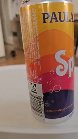 The video shows a can of Paulaner Spezi Zero on a white surface. The camera moves around the can, providing a close-up view of its design, which includes yellow, orange, red, and purple sections, as well as the "Paulaner" and "Spezi" logos. "Zero" is printed on the top of the can. The background is blurred, but shows elements of a domestic interior, including furniture and a scooter. The focus remains on the can of Spezi Zero.