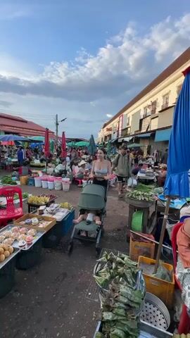 A woman is pushing a stroller with a baby in it through a busy outdoor market. The market appears to be selling produce and other goods. There are many stalls with umbrellas and awnings, and people are browsing and shopping. The woman is wearing a grey tank top and black shorts. The baby is wearing a diaper. The sky is cloudy.