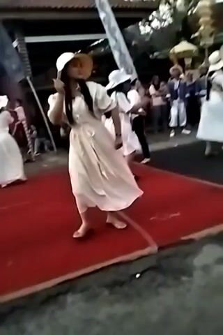 A group of young women wearing white dresses and straw hats are dancing on a red carpet outdoors. They appear to be performing a traditional dance. In the background, there are buildings, trees, and other people watching the performance. The sky is visible, suggesting it is daytime. The overall atmosphere seems festive.