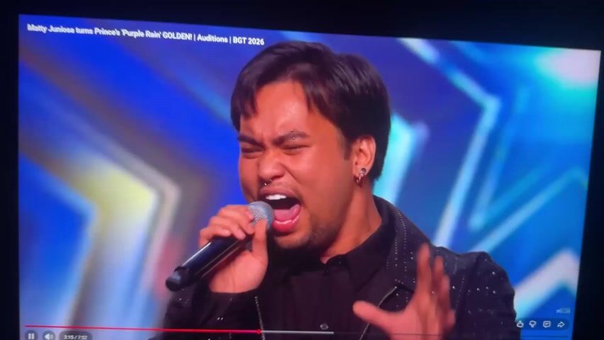 A man with dark hair is singing into a microphone. He has an eyebrow piercing and a nose ring. He is wearing a black shirt and a black jacket. The background is a blue and gold star pattern. The video is from a show called "Britain's Got Talent". The title of the video is "Matty Juniosa turns Prince 'Purple Rain' GOLDEN! Auditions | BGT 2026".