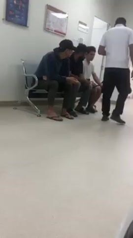 The video shows a waiting area, likely in a medical facility. Three young men are sitting on a metal bench with a silver frame. The man on the left wears a dark hooded sweatshirt, green pants, and sandals. The man in the middle wears a black t-shirt and grey pants. The man on the right wears a white t-shirt and dark shorts. A man in a white shirt and black pants stands near a door and appears to be speaking with the seated men. The room has white walls and a light-colored, speckled floor. A medical poster is visible on the wall to the left. There's another framed picture on the wall, but the content is unclear. A door is visible in the background.