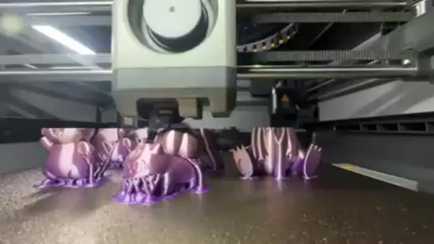 A 3D printer is shown up close, printing multiple small, purple objects that resemble cartoon characters. The printing head is a light gray color, and the printing platform is dark. The printer is contained within a gray and black casing, with bright overhead lighting. The printer is in operation.