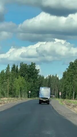 The video shows a black asphalt road with a white line down the center. The road is surrounded by trees on both sides. The sky is blue with white clouds. In the distance, a white truck is driving away from the camera. The truck is covered with a blue tarp. There are red and white circular signs on the side of the road. The video appears to be filmed from inside a vehicle, as the windshield wipers are visible in the foreground.