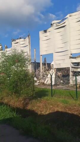 The video shows the exterior of a damaged building, possibly a warehouse or industrial facility. The building's facade is covered with large, white tarps that are ripped and torn, revealing structural damage underneath. Pink rectangular elements are visible on some of the tarps. There is a fence in front of the building with green posts. Vegetation, including grass and a small tree, grows in front of the fence. The sky is blue with scattered clouds. The video appears to be taken from a moving vehicle, possibly a car, as the lower portion of the frame shows part of a car's interior, including a dashboard and a side mirror. Some debris is visible on the ground near the building, and a car can be seen in the background behind the fence. The overall scene suggests significant structural damage, possibly due to a fire or explosion.