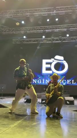 A video shows two men performing on a stage. The man on the left, wearing a tropical-print shirt and white pants, plays a saxophone. He stands with his left leg forward and his right leg back, leaning into his instrument. The man on the right, wearing a hat, sunglasses, and a khaki shirt, kneels on his right knee and plays a guitar. He holds the guitar across his body and gestures with his left hand. In the background, a large screen displays a logo featuring "EO" with a hat and the text "NGİN CELE." Stage lights are visible overhead.