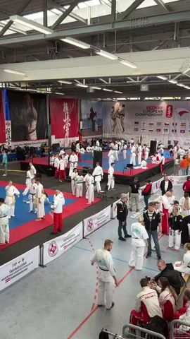 This video captures an indoor martial arts event, likely the 37th KWF European Championship, as indicated by a prominent banner. The setting is a brightly lit sports hall, where numerous individuals, predominantly adults and some younger participants, are gathered. Most people are dressed in traditional white karate gis (uniforms), while others wear red and white tracksuits or casual clothing.

Multiple mat areas, colored red and blue, are set up on the floor. On these mats, participants are seen warming up, practicing techniques, or preparing for competition. Others observe from the sidelines, some sitting on chairs or standing.

Large banners adorn the walls, providing contextual information. One large banner clearly displays "37th KWF EUROPEAN CHAMPIONSHIP" alongside the flags of various European countries including Armenia, Georgia, Poland, England, Denmark, Sweden, Norway, Russia, and Hungary. Another banner, located closer to the ground, reads "ŚWINOUJŚCIA AKADEMIA KARATE KYOKUSHIN" (Swinoujscie Kyokushin Karate Academy), suggesting an organizing or host entity from Poland. Two large speakers and several chairs are also visible within the venue. The overall scene depicts a busy and organized karate competition or large-scale training event.