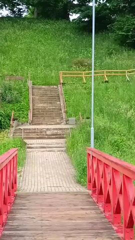 The video shows a park with a red bridge in the foreground and a grassy hill with stairs leading up. There are trees and bushes, as well as some benches on the hill. A lamppost is also visible. The scene appears to be a park or garden area.