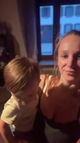 A medium shot shows a blonde woman holding a toddler on her left hip. The woman is looking towards the camera and smiling. She is wearing a black tank top with lace trim. The toddler, wearing a yellow shirt and blue pants, is looking down and to the left. The background is dimly lit and appears to be an indoor setting, possibly a living room, with furniture and a window visible. The video is taken from a first-person perspective, as if the woman is recording herself. The timestamp indicates the video is just a few seconds long.