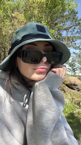 A young woman is featured in this video, enjoying a sunny day outdoors by a body of water, likely a river or lake, in Sneznevo, Russia. She is wearing a green bucket hat with a black band and a logo on the front, black rectangular sunglasses, and a grey hooded sweatshirt.

The video shows her in various relaxed poses. Initially, she is sitting upright, looking towards the camera with a slight smile. The background reveals a wide river, lush green hills, and some power lines on the distant hills. Pine trees are visible near her.

In the later shots, she lies down on a patterned blanket or mat on the grassy bank. Her dark hair is visible beneath the hat. She continues to look at the camera, and in the final frame, she raises her hand to form a peace sign. The weather is clear and bright, indicating a pleasant sunny day.