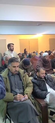 The video shows a gathering of mostly middle-aged men in a large room. Most of the men are sitting in wooden chairs and facing towards the front of the room, where a man in a light-colored blazer is standing. Some men are taking notes. There is a woman in the background wearing a black hijab. A few people in the back are holding cameras and recording the event. The room is well-lit with a white ceiling and light-colored walls. There appear to be some people standing behind the chairs and near the back of the room. The overall atmosphere is attentive and formal.