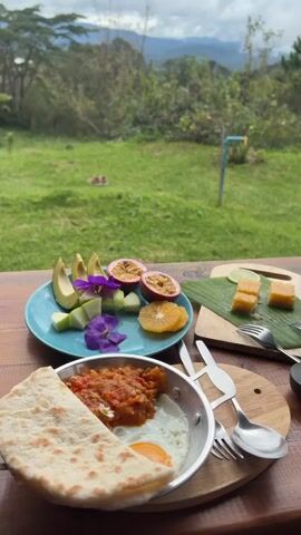 A vertical video displays a breakfast spread on a wooden table with a lush green landscape and mountains in the background. On the table, there's a plate of sliced avocado, cucumber, orange, and passion fruit, a small pan with a fried egg and tomato salsa, flatbread, a wooden cutting board with small cubes of a yellowish dessert and lime, a small teapot with tea, a tiny teacup, a stack of two books, and cutlery. The camera pans to the right, focusing on the teapot, books, and the background scenery. The sky is cloudy.