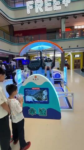 The video shows a young boy in a VR paraglider simulator inside of a shopping mall. The boy is seated in a swing suspended from a frame shaped like a paraglider wing. He is wearing a VR headset and holding the handles. To the left, another young boy is standing with an adult, both looking at the simulator. The simulator has a screen that displays the VR paragliding experience. A sign at the base says "VR Paraglider". Behind the simulator, there are other arcade games and a neon sign that reads "FREE". The shopping mall environment is visible in the background, including other stores and walkways.