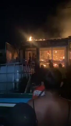 The video shows a building engulfed in flames at night. People are observing the fire, some are gesturing towards it and shouting. The structure appears to be a one-story building with windows, and signage that reads "MaUka" is visible on the roof. Smoke is billowing into the night sky. The perspective is from a slightly elevated position, possibly from a vehicle or a raised platform. A large blue container is in the foreground, partially obstructing the view.
