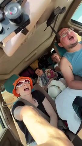 Four people are inside a packed car, taking a selfie video. The camera is positioned to capture the driver in the front left seat, a woman wearing an orange hat and sunglasses, and her right arm is extended, holding the camera. In the middle row, a woman in a floral shawl and a white hat is seated behind the driver. Next to her, a woman with blonde hair smiles. In the passenger seat, a man wearing glasses and a blue shirt is leaning back and appears to be speaking. The car's interior is filled with luggage, including several large suitcases stacked behind the passengers. The environment outside the car appears to be a dry, hilly landscape under a bright sky, suggesting a road trip in a warm climate. The video is set to music.