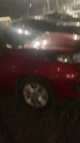 The video captures a row of parked cars at night in Angeles, Philippines. The scene is dimly lit, with overhead lights casting reflections on the vehicles. In the foreground, the hood and headlight of a shiny red car are prominent. Behind it, several other cars are visible, mostly dark in color, parked on a gravel surface. The perspective suggests the video was taken from within a car or close to the ground. The lighting and darkness indicate it is nighttime. There are no people visible in the frame. The background shows the silhouettes of structures and possibly more vehicles, suggesting a parking lot or an open area.