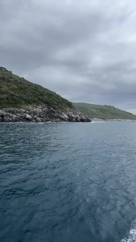 The video shows a body of water next to rocky, grassy hills under a cloudy sky. The water is dark blue and slightly choppy. The hills are green and appear to be covered in vegetation. The sky is overcast with grey clouds. The video appears to be taken from a boat or ship.